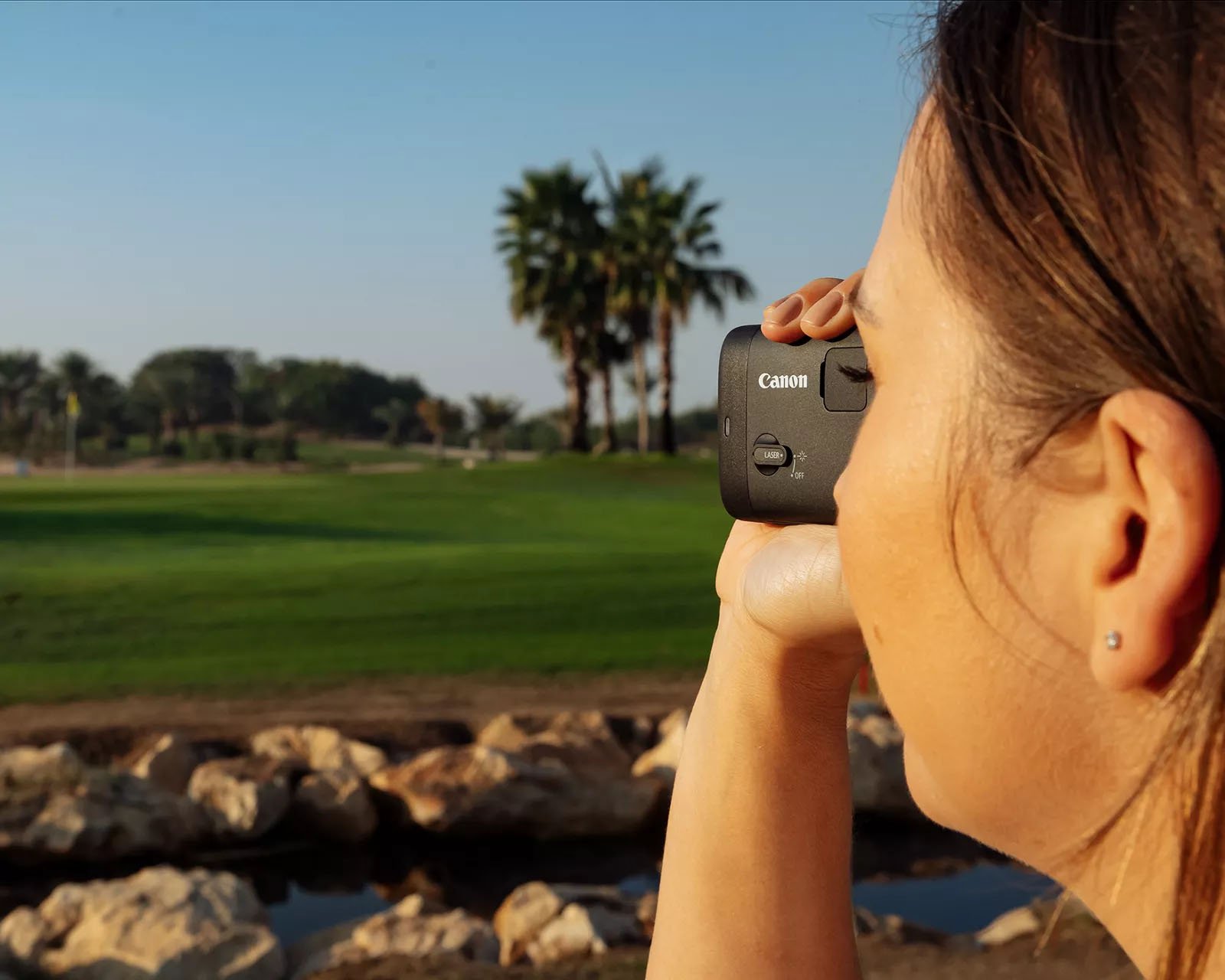 A person with long hair is holding a Canon rangefinder with an outdoor landscape featuring a green field, rocks, and distant palm trees under a clear blue sky. The picture is taken from the side, showing the person focusing on something in the distance.