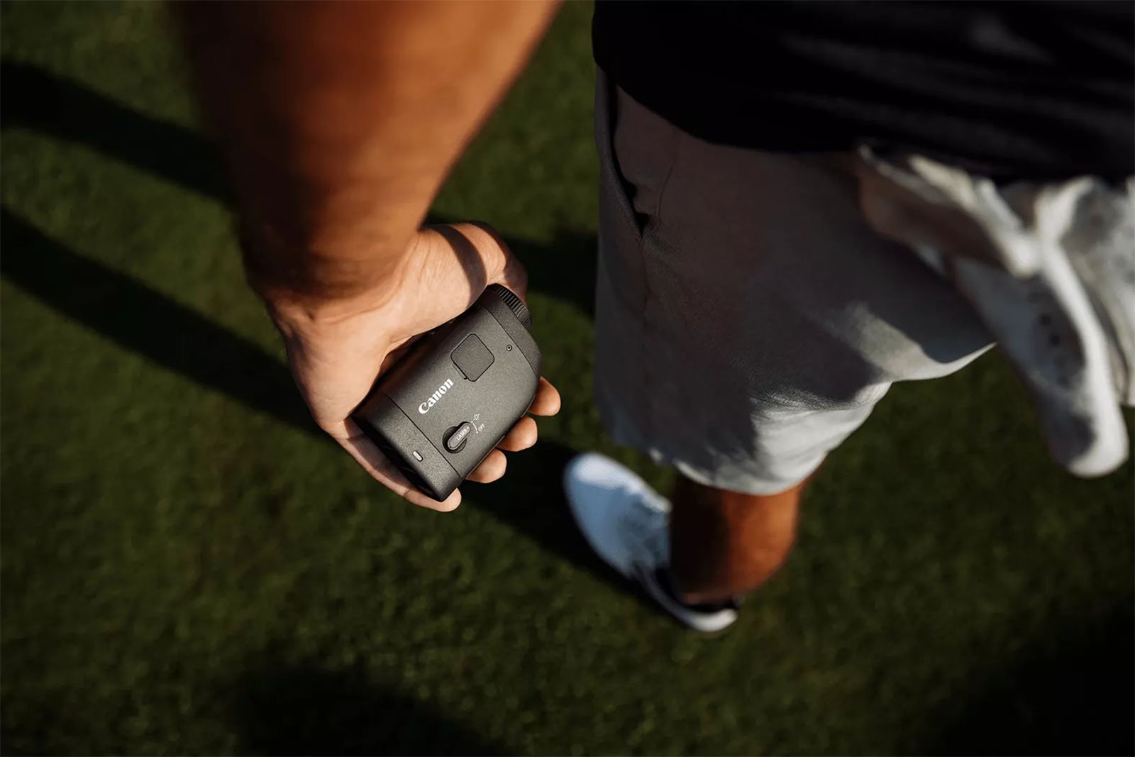 A person holds a Canon rangefinder in their right hand while standing on grass. The individual is wearing a black shirt, light-colored shorts, and white athletic shoes. The perspective is from their point of view looking down at the rangefinder.