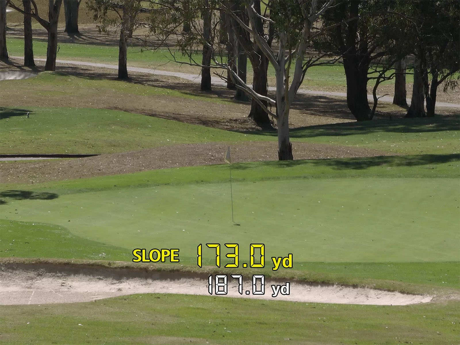 A golf course view featuring a green surrounded by trees. A yellow text overlay indicates "SLOPE" with a numerical value of "173.0 yd," and below it in white text, "187.0 yd" is shown.
