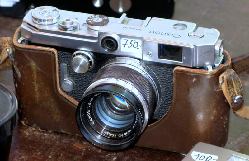 A vintage Canon film camera with a leather case is displayed on a surface. The camera has various dials and a large lens marked "Canon Lens." A small sticker on the camera reads "750." Another sticker in the bottom right corner reads "100.