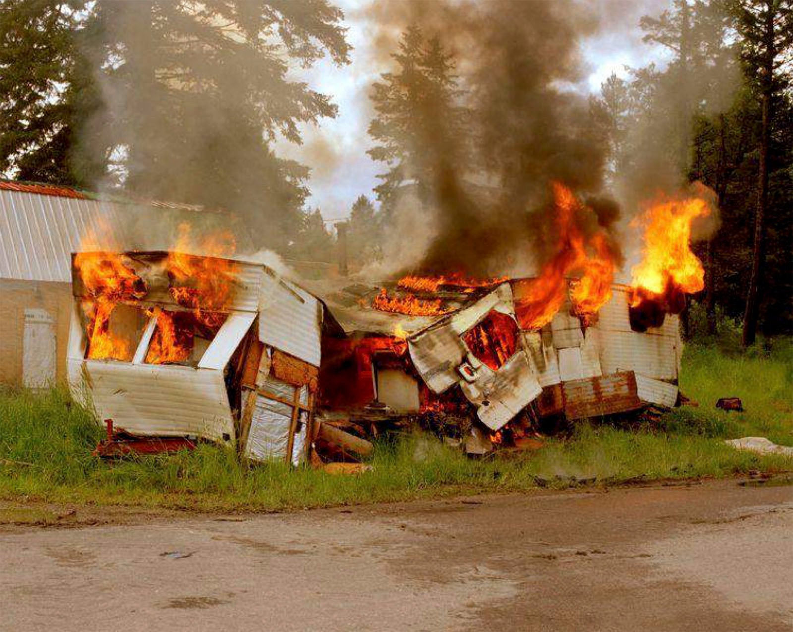 A dilapidated mobile home is engulfed in flames next to a forest, emitting thick smoke under a cloudy sky. the structure is mostly destroyed with visible bright orange and yellow fire consuming it.