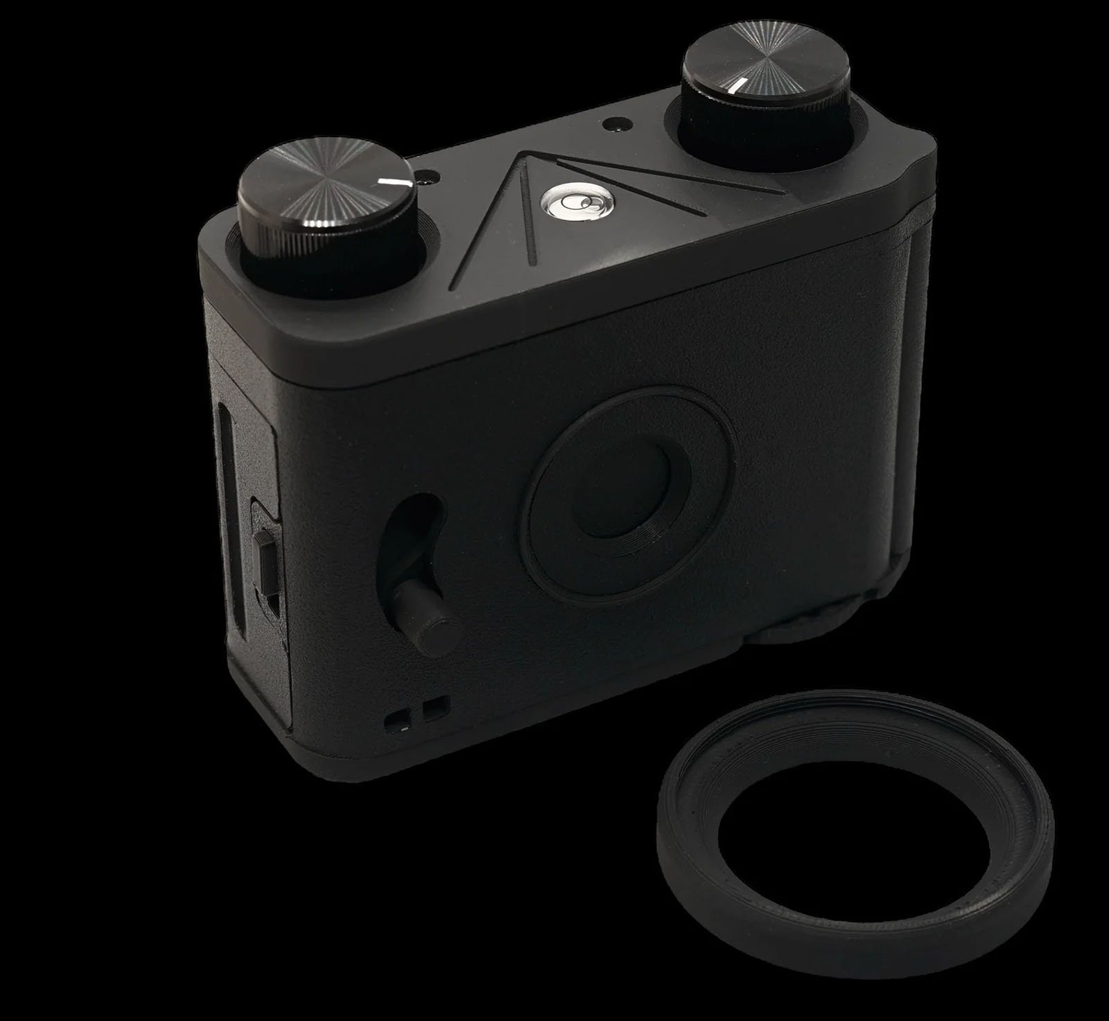 A compact black camera with two large dials on top and additional controls on the sides. It has a minimalist design with a circular element on the back. A separate lens or attachment piece lies beside it against a black background.