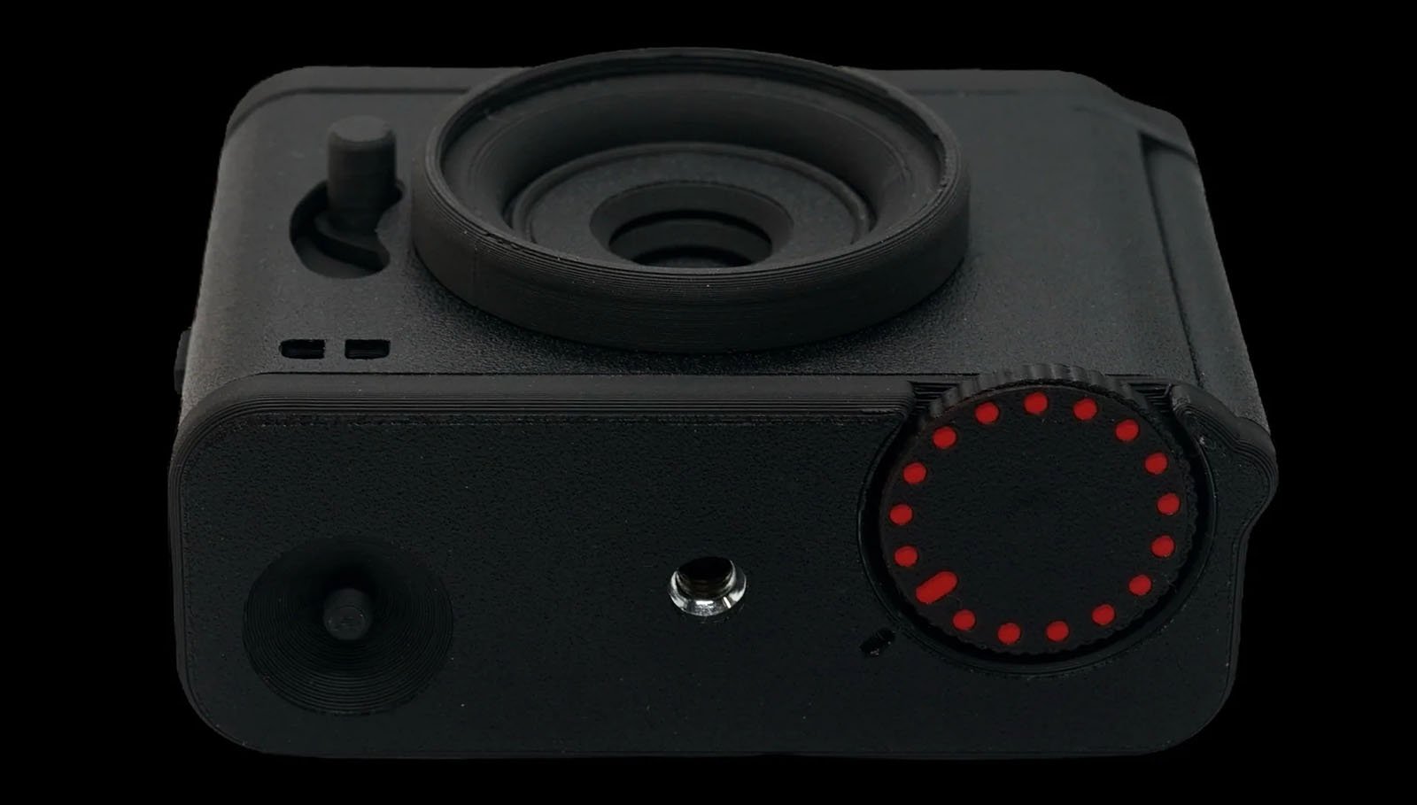 A close-up of a black, compact camera with a large lens in the middle. There's a circular control dial with red markers on the right side and a small protruding button towards the top left. The camera has a minimalist design against a black background.