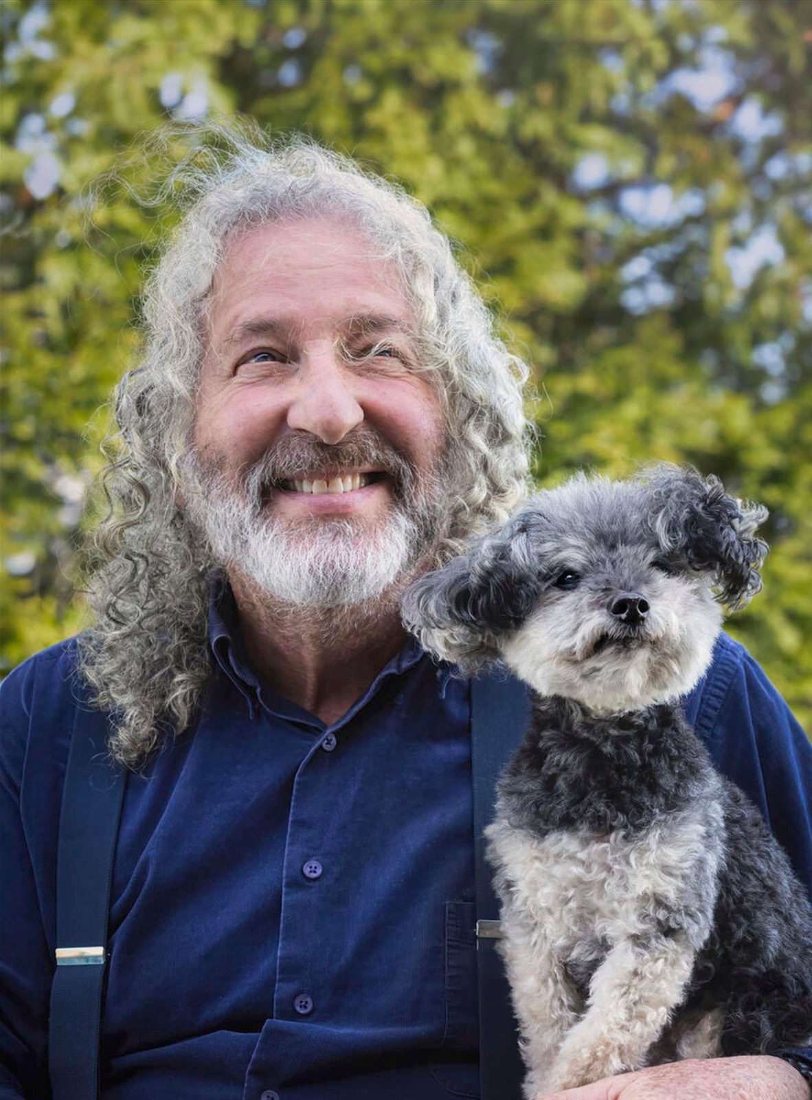 A cheerful elderly man with curly gray hair holding a small black and white dog, both smiling at the camera, against a blurred green foliage background.