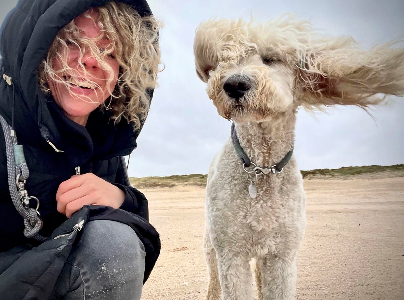 A joyful moment on a windy beach with a woman and a fluffy dog, both with hair blown by the wind, smiling and enjoying the breezy weather together.