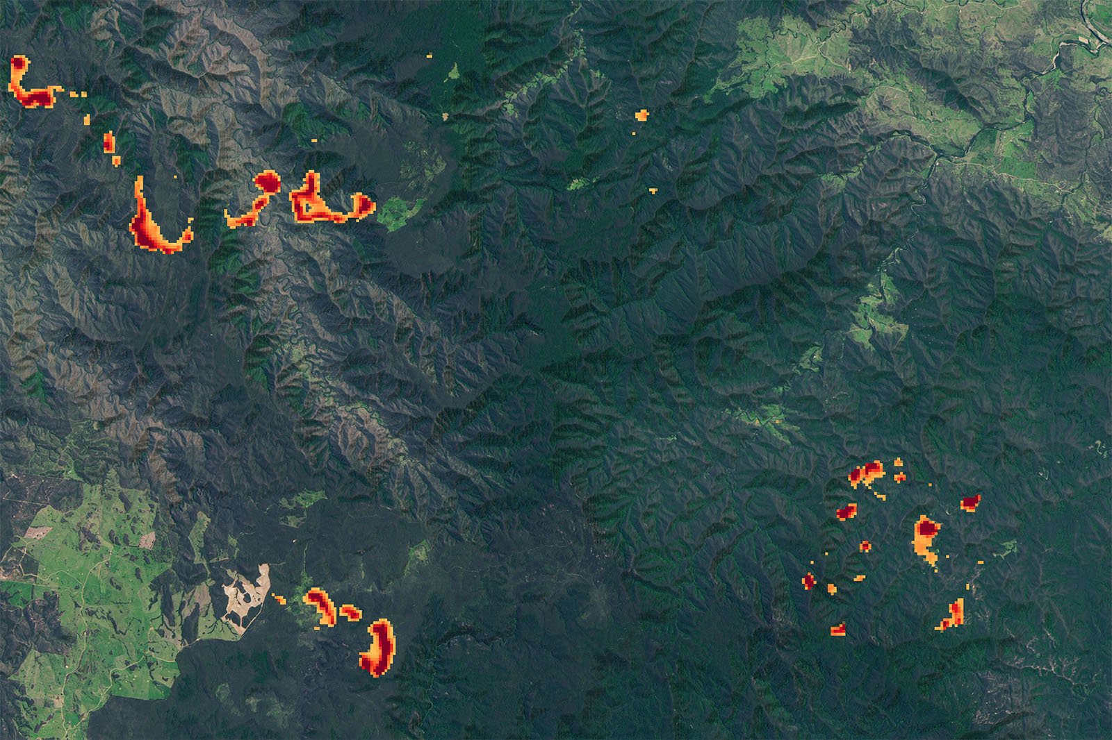 A satellite image showing a mountainous area with wildfires burning. Bright red and yellow areas indicating active fire zones are scattered across the landscape, especially on the left side and bottom right corner, contrasting with the surrounding green terrain.
