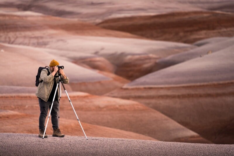 A photographer stands on a rocky terrain, using a tripod to steady their camera. The backdrop features smooth, rolling hills in shades of brown and gray. The photographer is dressed warmly, wearing a beanie and a backpack, focused on capturing the landscape.