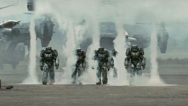 Four armored robots emerge from a smoky, mist-covered area on a desolate ground. In the background, large futuristic aircraft hover above the ground, contributing to the haze. The scene appears intense and action-packed.