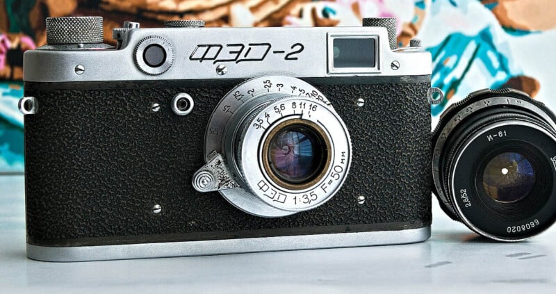 A vintage FED-2 rangefinder camera with a textured black and silver finish is placed on a white surface. Next to it, on the right, is a detached camera lens. The background features a colorful, blurred painting.