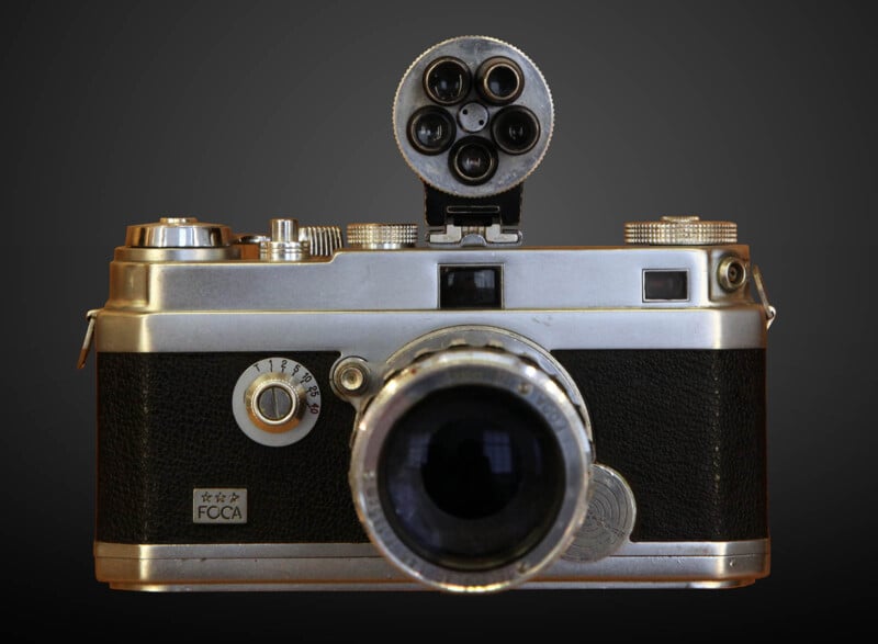 A vintage, silver and black rangefinder camera with a prominent lens in the center and several dials and knobs on the top. It has a unique four-lens viewfinder attachment on top. The camera is displayed against a dark gradient background.