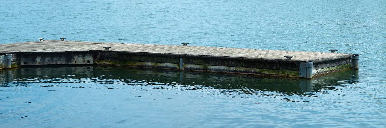 A wooden pier with metal cleats floats on calm blue water, showing signs of weathering and algae accumulation along its submerged edges.