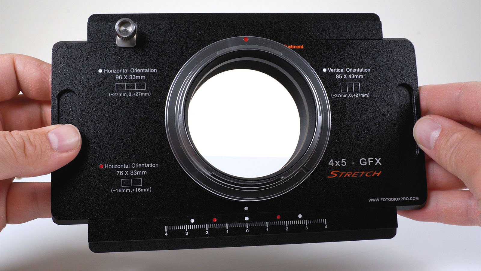 Two hands holding a fotodiox gfx stretch adapter, which enables the use of large format lenses on medium format cameras. the black adapter is displayed with measurement markings and orientation labels.