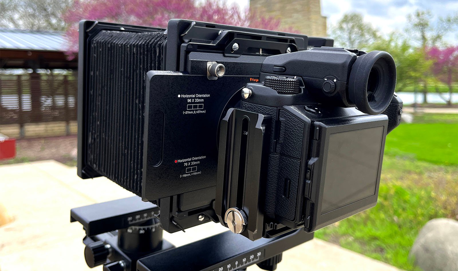 A close-up of a large format camera mounted on a tripod outdoors, focusing on its intricate black body and controls, with a blurred green park background.