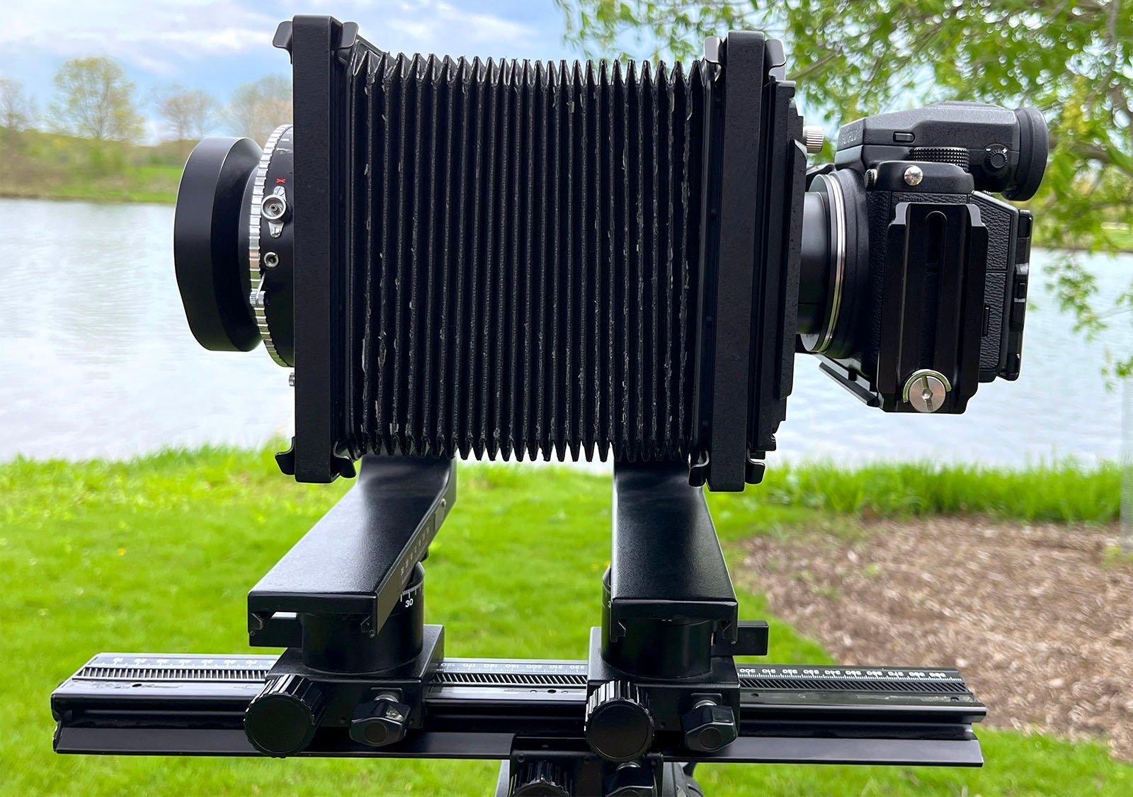 A large format camera mounted on a tripod, positioned outdoors with a scenic lake and lush trees in the background. the bellows of the camera are fully extended.