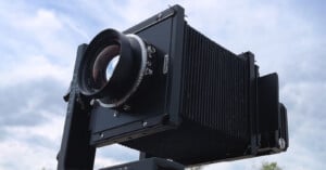 A large format camera with a bellows focusing system is positioned against a cloudy sky, emphasizing its lens and intricate design details.