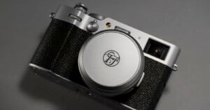 A silver and black Fujifilm camera lies on a gray surface. The camera features a retro design with a round lens cap displaying the Fujifilm logo. The body has a textured grip and various control dials visible on the top.