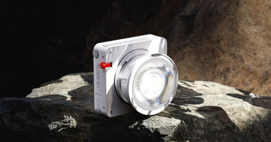 A modern white digital camera labeled "ML100" with a large lens is positioned on a rocky surface, partially illuminated by sunlight. The background consists of uneven, natural rock formations.