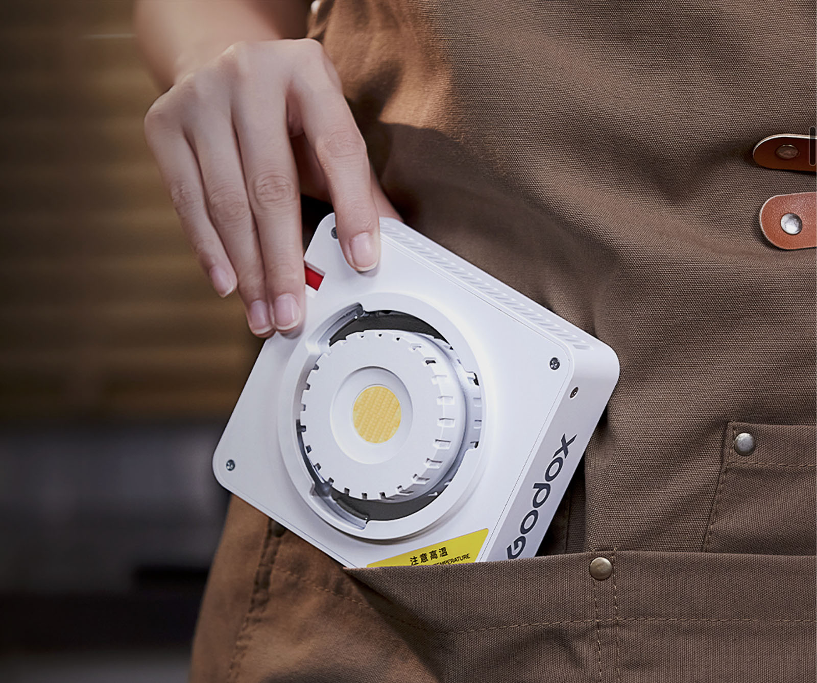 A person wearing a brown apron is holding a compact white device with a circular component and text on it. The device is partially tucked into the apron pocket. The background is blurred, focusing on the device and hand.