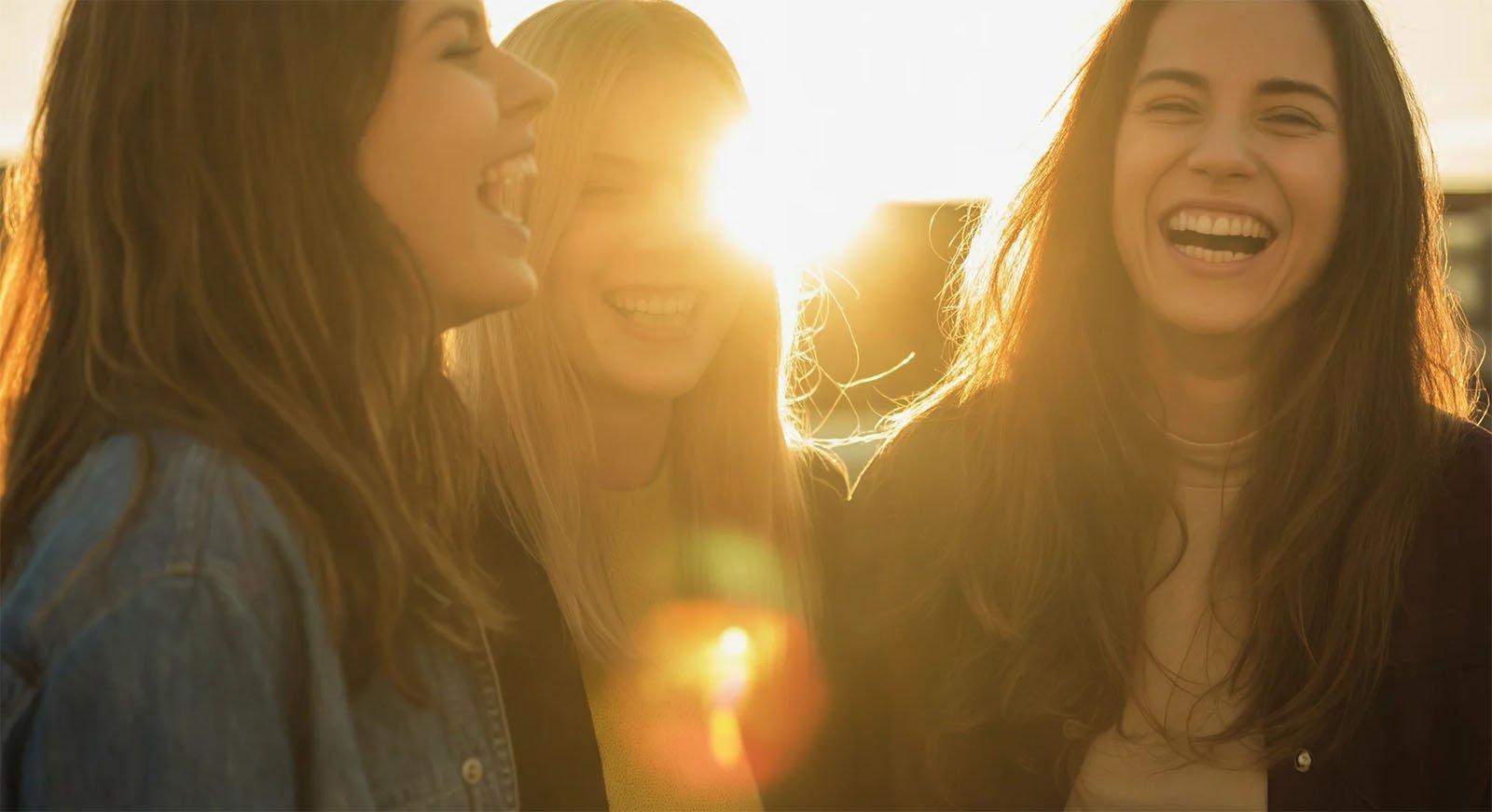 Three women are laughing and standing close together in the foreground, illuminated by warm sunlight from a low horizon behind them. The light creates a lens flare effect, highlighting their joyful expressions. They appear to be outdoors during sunset.