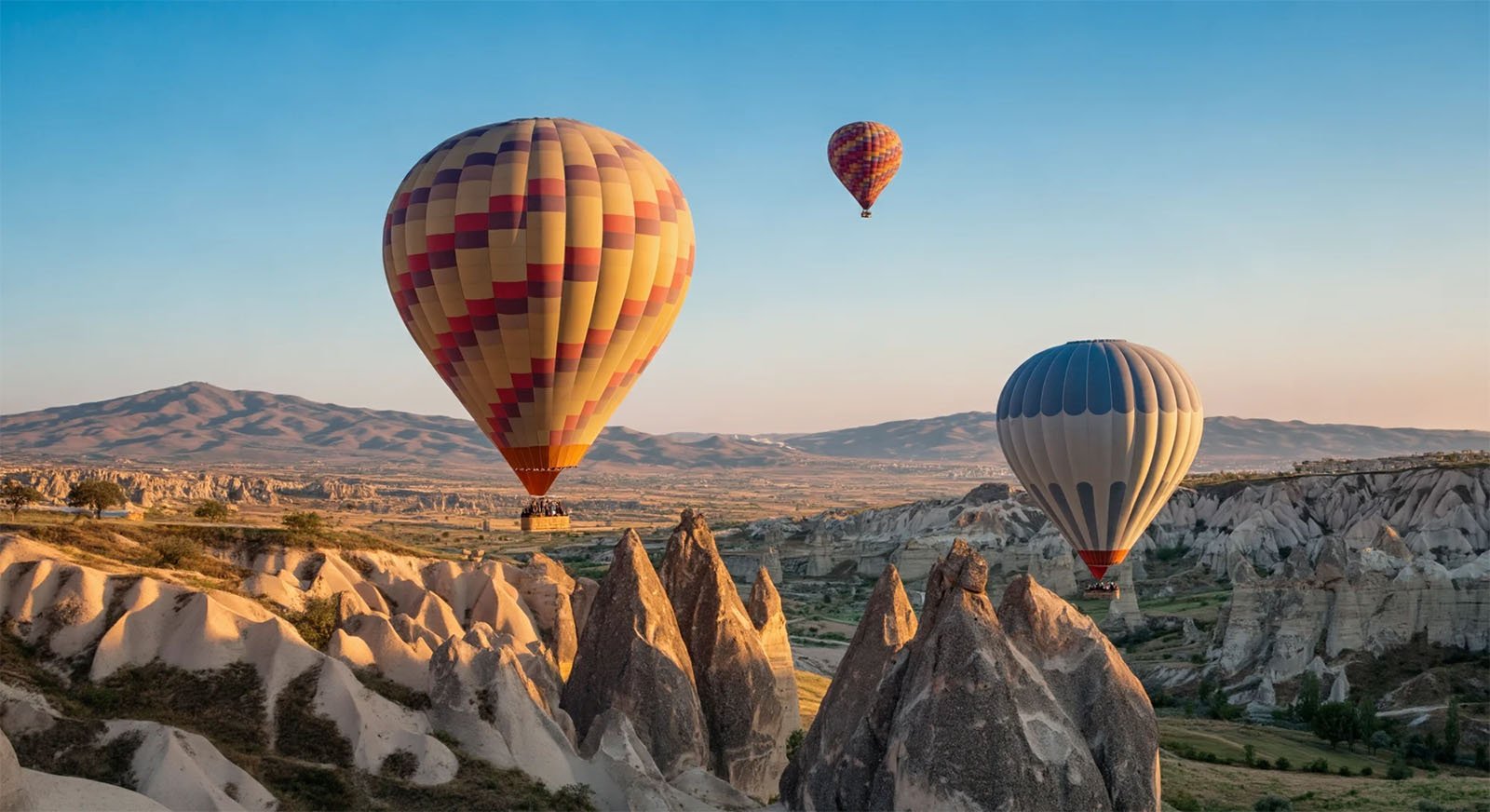 A picturesque landscape featuring three colorful hot air balloons floating over unique, rocky formations during sunrise or sunset. The sky is clear, and the distant mountains frame the scene, adding to the serene and captivating view.