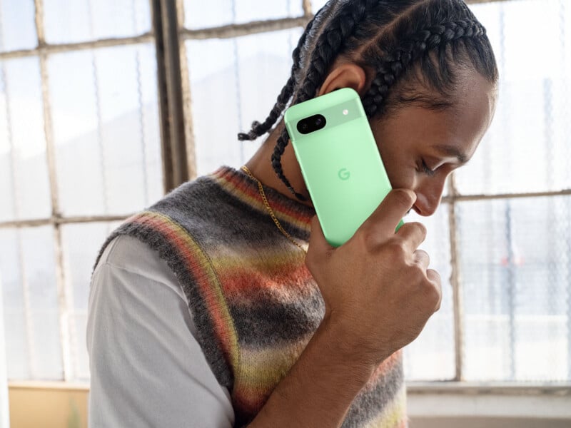 A man with braided hair talks on a Google Pixel 8a smartphone in the color Aloe. He's indoors by a window with grid-like panes wearing a colorful knitted sweater.