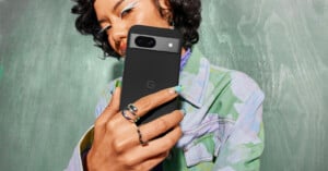 A woman with curly hair taking a selfie with a Google Pixel 8a phone, focusing on her stylish rings and green nail polish, against a green textured background.