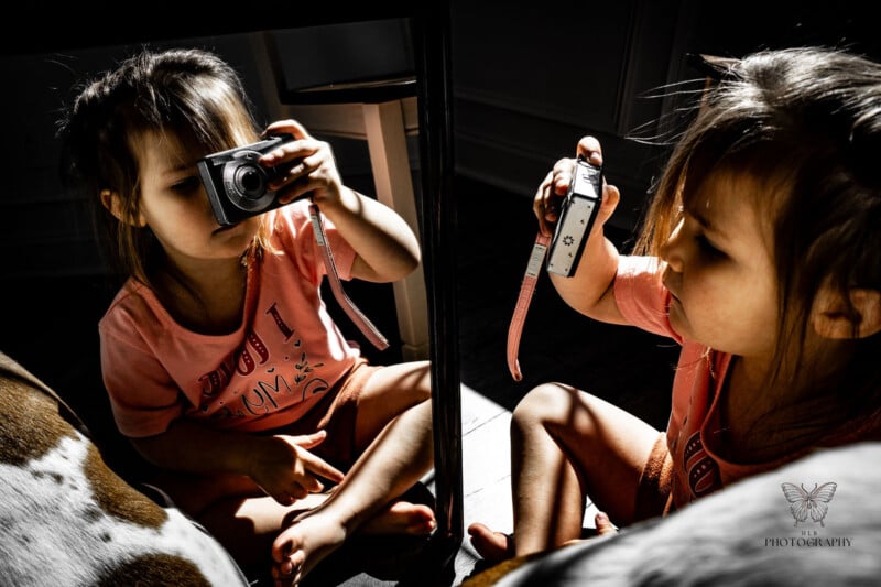 A young girl in a pink shirt holds a compact camera, taking a photo of her reflection in a mirror while sitting in a sunlit room.