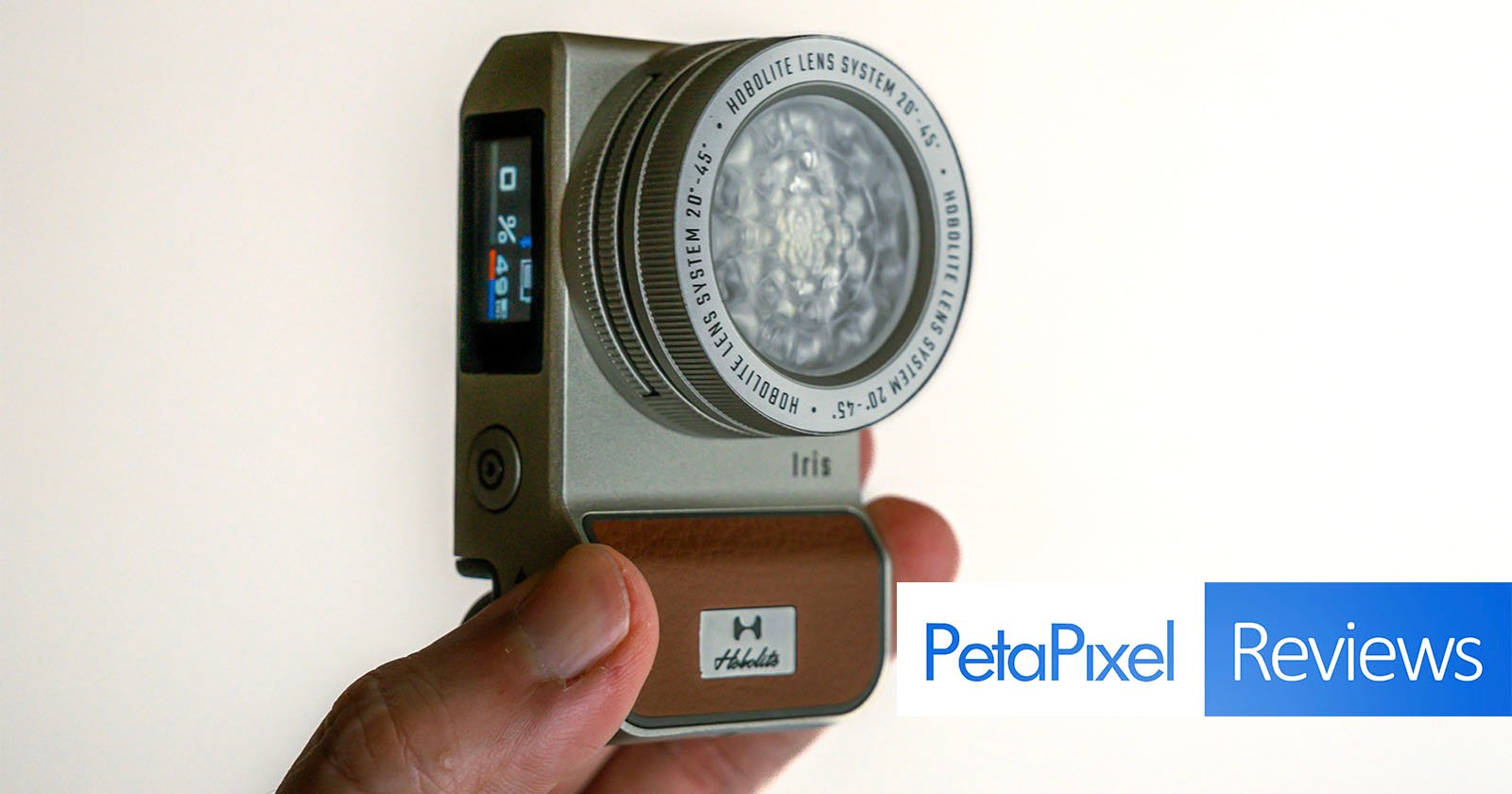 A hand holds an Iris Hobalux camera. The camera features a round Noblige Lens System and a small screen on the side. The bottom of the camera has a brown panel with the Hobalux logo. The text PetaPixel Reviews is overlaid in the lower right corner.