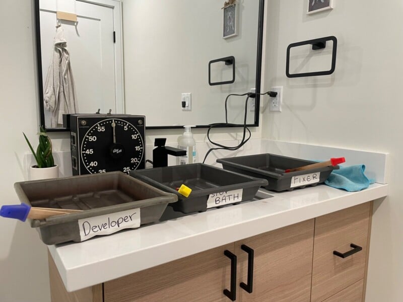 A modern bathroom setup converted into a darkroom with three labeled trays on the counter for film development: "developer," "bath," and "fixer." accessories like a clock, tongs, and a towel are also visible.