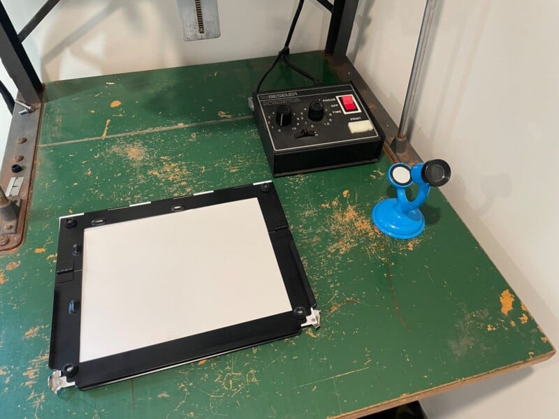 A screen printing station with a blank frame on a stained table, accompanied by an exposure light meter and a small blue suction cup tool.