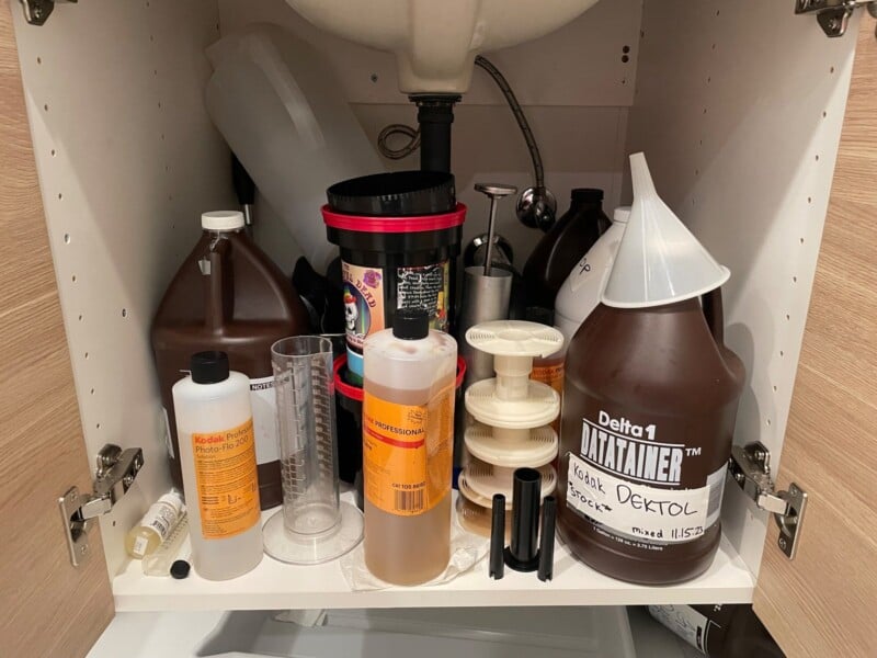 A cluttered cabinet filled with various darkroom supplies including chemicals, film developer bottles, and measuring equipment used for photographic processing.