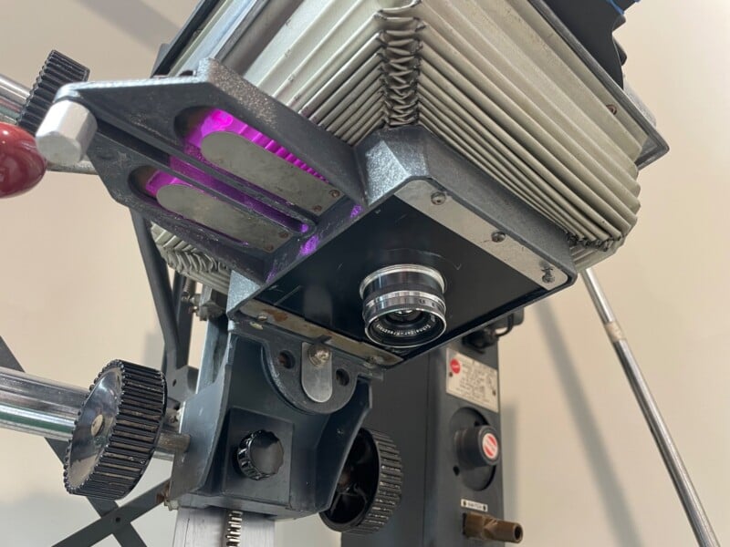 Close-up view of a vintage movie projector showing its lens, gears, and light system, with detailed elements of metal housing and adjustment knobs.