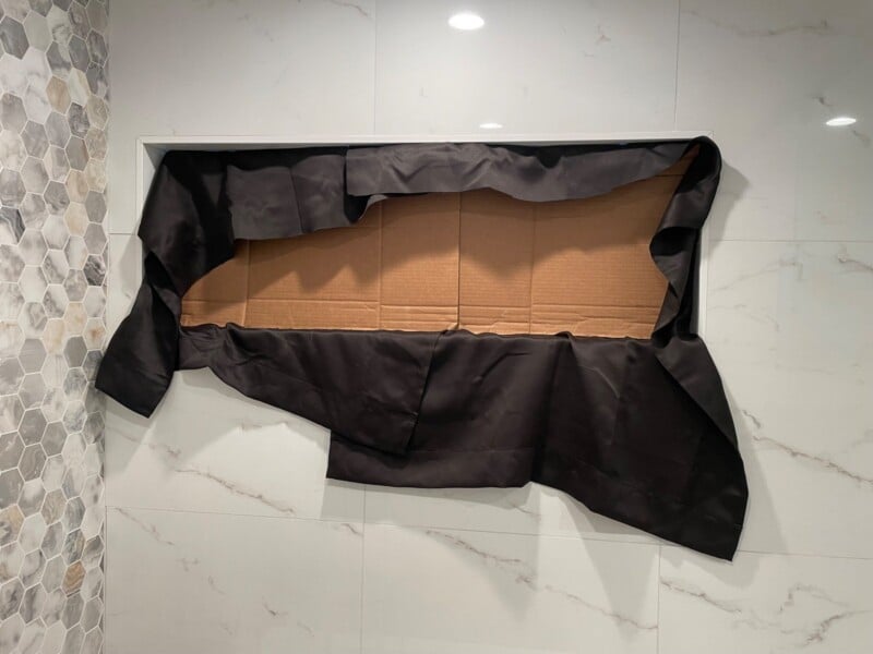 A covered rectangular niche in a marble wall with a black tarp secured over it, partially exposing the cardboard underneath. the surrounding wall features hexagonal and marble tile designs.