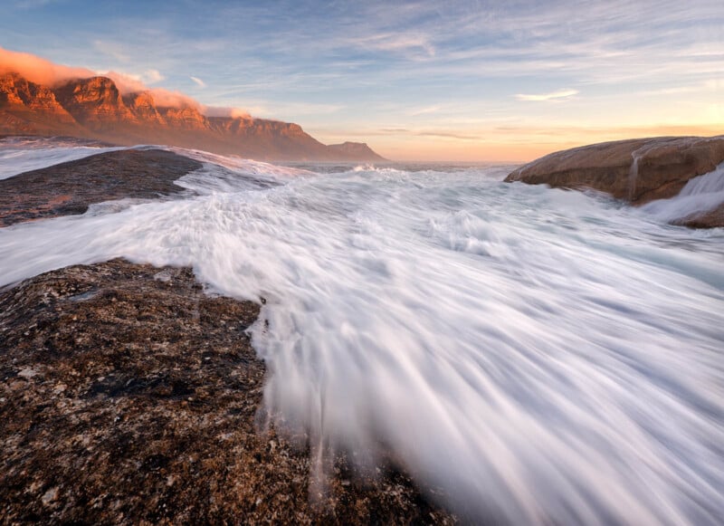 Rocky shoreline with waves crashing over the rocks, creating a dynamic flow of white water. In the background, a mountain range is illuminated by the soft glow of a setting sun, with a partly cloudy sky adding to the serene yet dramatic landscape.