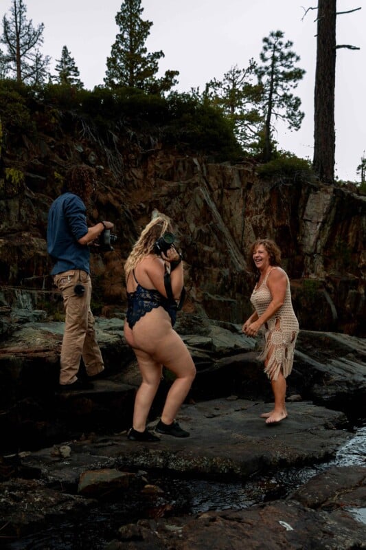 Three people are on rocky terrain in front of a cliff. Two are photographers, one in casual attire and another in a blue one-piece swimsuit, both taking photos of the third person. The third person, wearing a beige crochet dress, is smiling and posing barefoot. Pine trees are visible in the background.