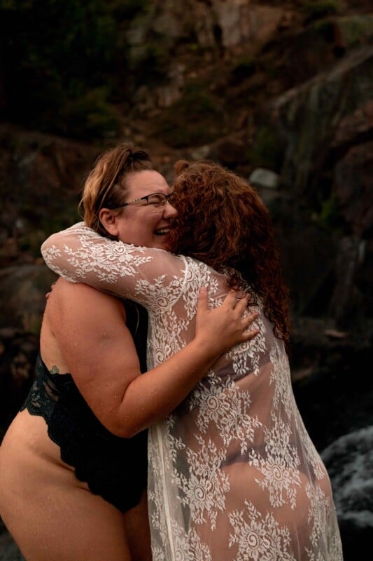 Two individuals embrace joyfully near a waterfall. One wears a black swimsuit, and the other wears a lacy white cover-up over a swimsuit. They are smiling broadly, sharing a moment of happiness and connection amidst a natural rocky backdrop.
