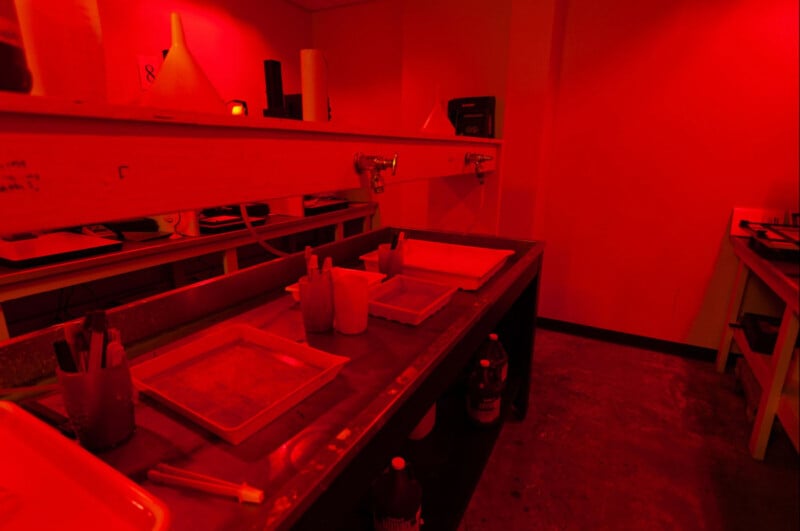 A darkroom illuminated in red light with photography development trays on benches, bottles of chemicals, and tools used for film processing.