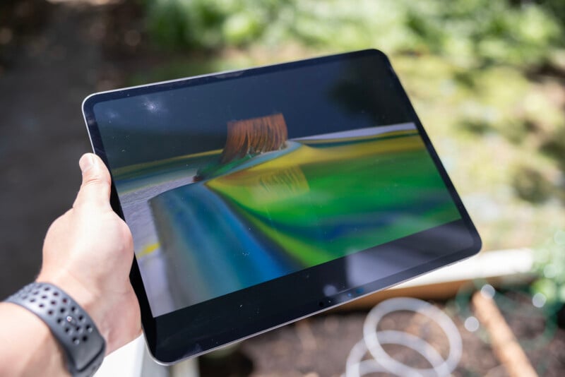 A hand holding a tablet displaying a colorful abstract image, outdoors with blurred greenery in the background.