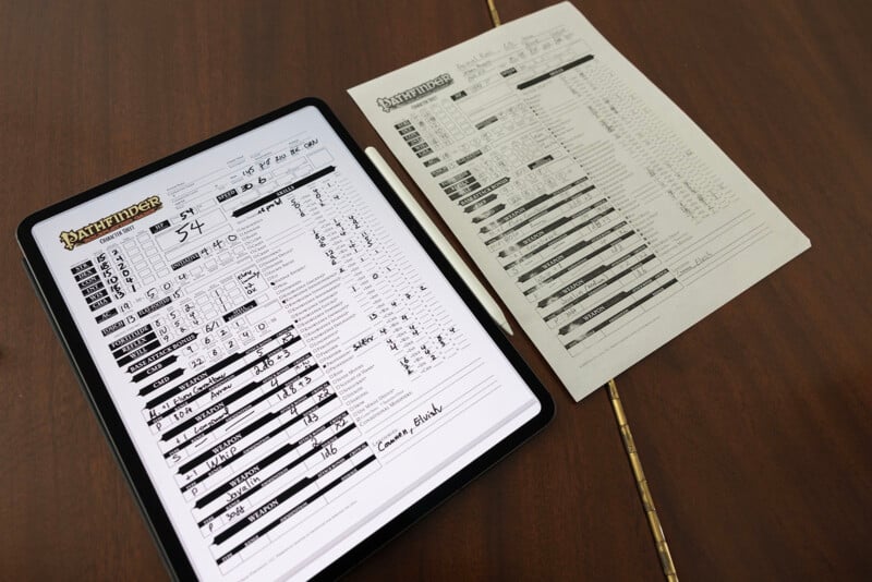 An iPad displaying a digital character sheet for the Pathfinder role-playing game next to a printed character sheet on a wooden table.