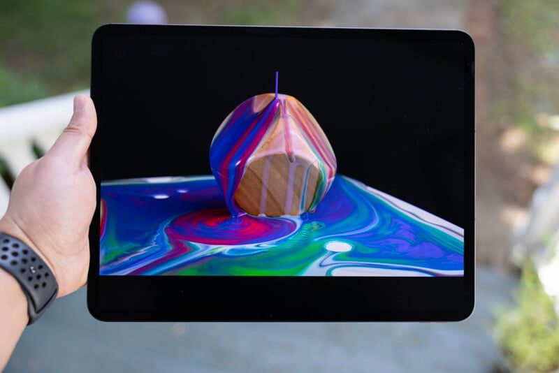 Hand holding a tablet displaying a 3D image of a colorful apple with striking ripple effects around it, symbolized in vivid blues, purples, and reds.