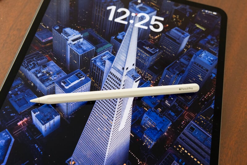 An iPad displays a cityscape wallpaper showing skyscrapers at night, overlaid with the time "12:25". An Apple Pencil rests diagonally across the screen.