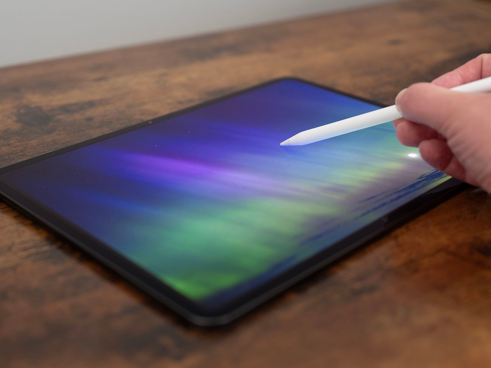 A hand holding a stylus hovers over a tablet screen displaying a colorful gradient of blues, purples, and greens, resembling the aurora borealis. The scene is set on a wooden surface.