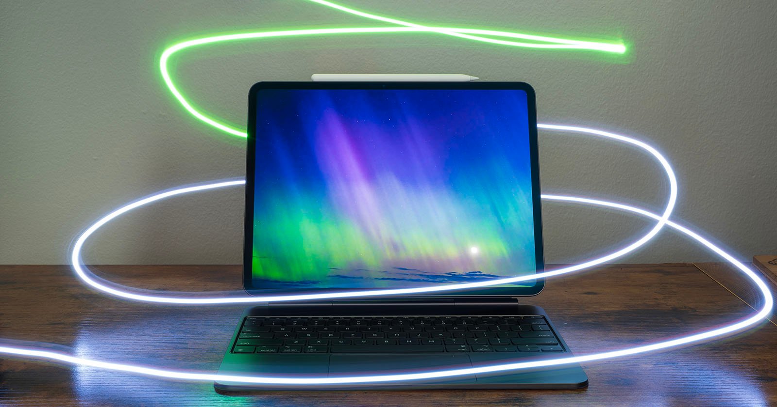 A tablet with a keyboard is shown on a wooden surface. The screen displays a colorful aurora image. A light pen rests on top of the tablet, and glowing light trails in green and white swirl around the tablet, creating a dynamic visual effect.