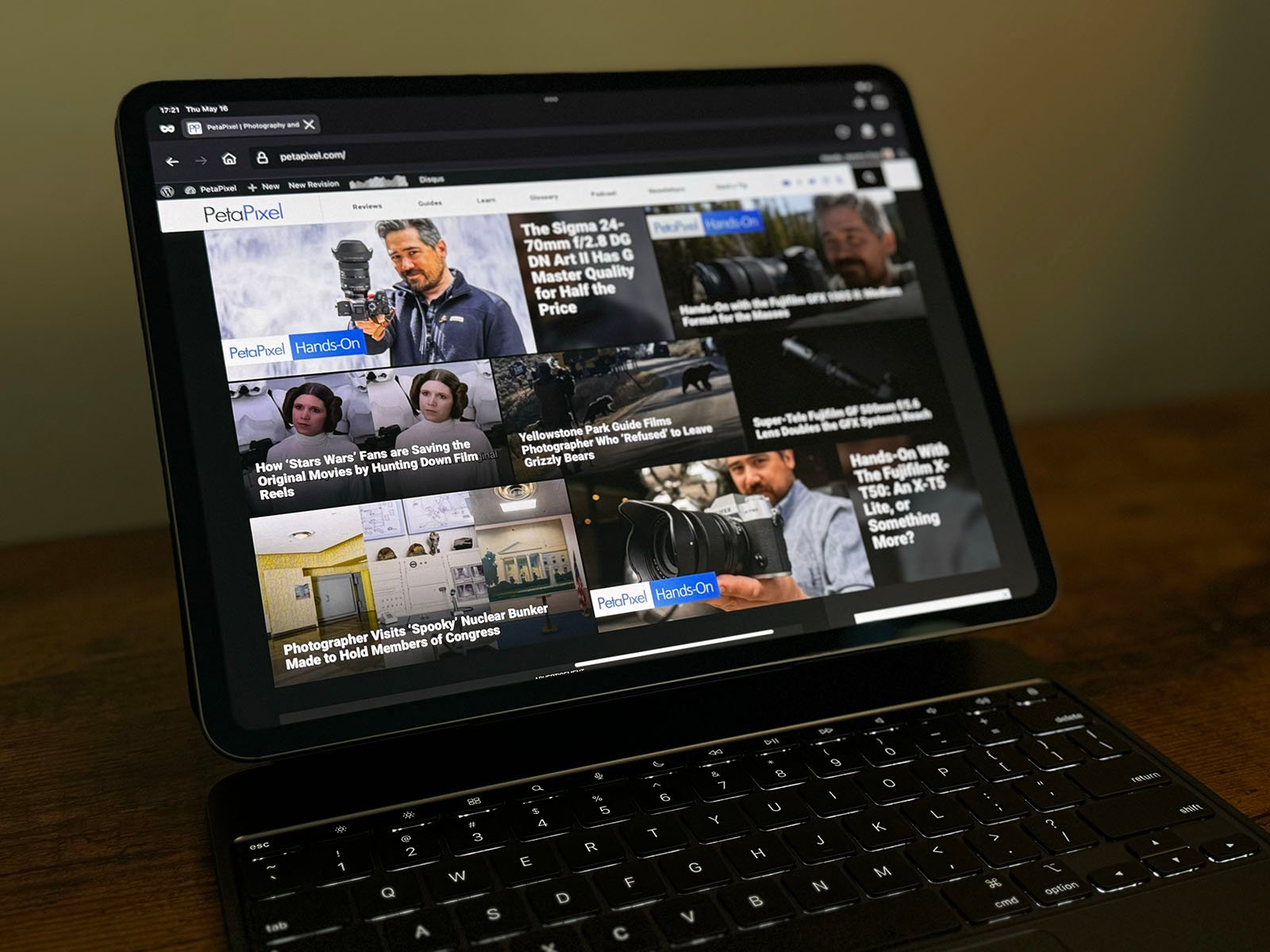 A tablet with an attached keyboard displays a website featuring various photography-related articles. The homepage shows multiple thumbnails of images and headlines, including topics on cameras, photography techniques, and notable photographers.
