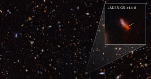 A deep field image of space showing numerous distant galaxies and stars. An inset zooms in on galaxy JADES-GS-z14-0, highlighted with an arrow, displaying a more detailed view of the small, red galaxy against the dark backdrop of space.