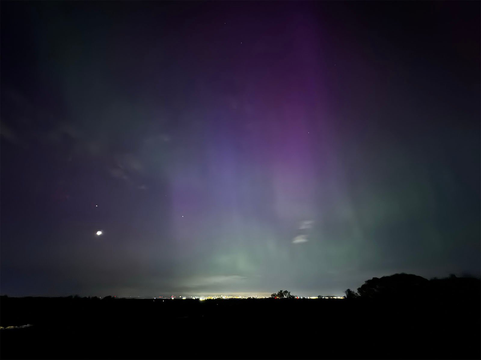 A night sky illuminated by a pale purple aurora with stars visible and a bright celestial body, possibly a planet or moon, shining vividly above a faintly lit landscape.