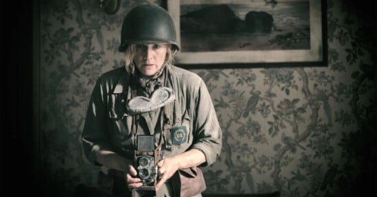 A woman in a vintage military uniform and helmet holds an old camera, standing in a room with floral wallpaper, displaying a serious expression.