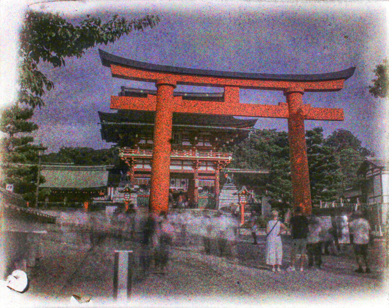 A grainy image of a vibrant red torii gate leading to a traditional japanese structure, surrounded by trees, with blurred figures of people in the foreground.