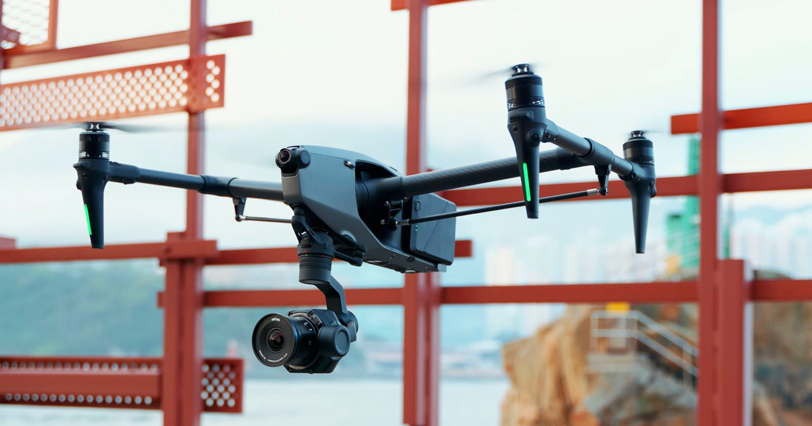 A black drone with a mounted camera flying by a red structure, with a blurred cityscape and water body in the background.