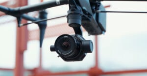 A close-up of a black camera mounted on a drone, with a blurred bridge structure in the background. the camera lens has a visible brand name.